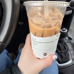 Frosted Caramel Latte - A hand with light skin and painted nails holds a large iced caramel latte in a clear plastic cup with a lid and straw. The cup has a white sleeve with a receipt attached, indicating it is a frosted caramel latte. The background shows the interior of a car, including the dashboard, air vents, and gear shift. The person is wearing a black jacket and blue jeans.