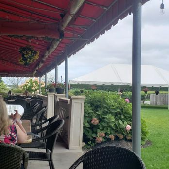 Clovis Point - An outdoor seating area with black chairs and a round table under a red canopy, overlooking a garden with bushes and a white tent. - Wineries Near Me - Jamesport, New York