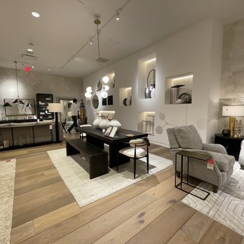 Arhaus - The image shows a furniture store interior with various living room furniture pieces, including sofas, armchairs, and tables. The space has wooden flooring, neutral-colored walls, and built-in wall niches with decorative items. Lighting fixtures illuminate the area, and there are customers browsing in the background. The overall style is modern and minimalist, emphasizing clean lines and neutral tones. - Home Decor Near Me - Troy, Michigan