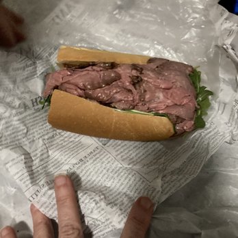 Bodega On Smith - Bodega Roast Beef sub - A roast beef sandwich with lettuce is wrapped in newspaper, placed on a plastic surface, with a person's hand pointing at it. - American Near Me - Providence, Rhode Island