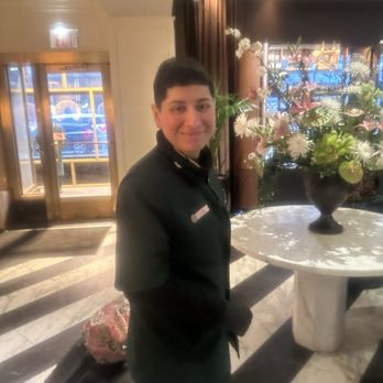 Pendry Chicago - Chris, door girl - A young hotel staff member with short dark hair, wearing a black uniform, stands in the lobby of a luxury hotel. The background features a large floral arrangement on a marble table, with natural light coming through the windows and a glass door leading outside. The lobby has marble flooring and decorative curtains, indicating an upscale setting. - Hotels Near Me - Chicago, Illinois