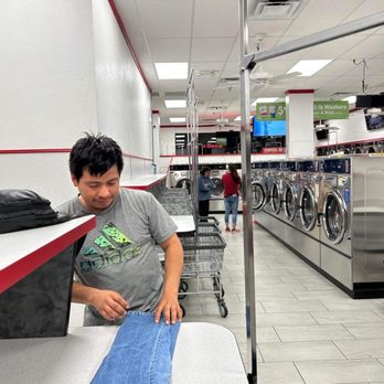 Coin Less Laundry - A person in a gray Adidas t-shirt is sitting at a laundromat, holding a pair of jeans. Laundry machines and other customers are visible in the background. - Laundromat Near Me - Phoenix, Arizona