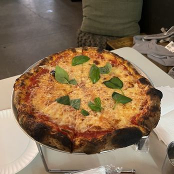 Farm.One - A round cheese pizza with melted cheese and tomato sauce is displayed on a metal stand on a table. Fresh basil leaves are scattered on top of the pizza. The table has a white surface with a paper napkin and a plastic-wrapped utensil nearby. In the background, there is a green cushion, some clothing, and a concrete floor, indicating an indoor dining environment. - Farms Near Me - Brooklyn, New York