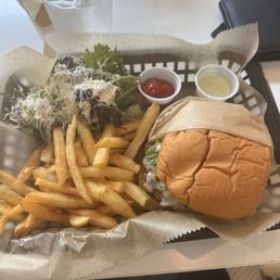 Sweet and spicy burger Daily Special - A tray contains a burger with a soft bun, fries, a small mixed salad with greens and sprouts, and two small cups of condiments—ketchup and a creamy sauce—on a white table at Mina Bistro in Miami, FL. A glass of water and a black wallet are also visible on the table.
