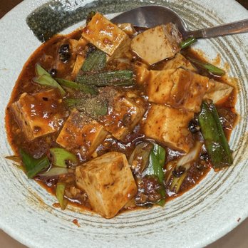 Too Hot - Stone Pot Tofu - A ceramic bowl holds a dish of tofu cubes coated in a dark, spicy sauce, garnished with chopped green onions and black pepper. A metal spoon rests in the bowl, and the dish appears to be part of Asian cuisine, likely featuring Sichuan or Yunnan flavors. - Asian Fusion Near Me - Cambridge, Massachusetts