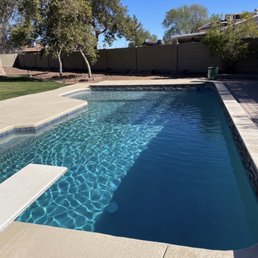 A backyard pool with clean, blue water, concrete decking, and trees in the background, enclosed by a privacy fence.