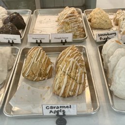 Sweet Scone - A bakery display case contains multiple trays of baked goods, primarily scones with various flavors such as vanilla bean, caramel apple, strawberry peach, and others. Each tray is labeled with the flavor name, and the scones are arranged in neat groups. The case has a glass front, allowing customers to view the selection of freshly baked scones and treats.