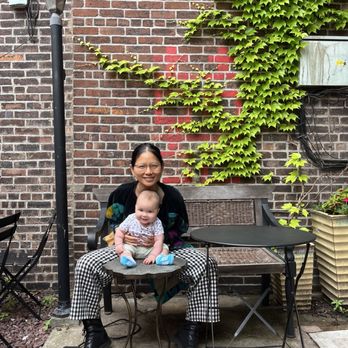 Red Rover - A woman with glasses and a baby sit on a small metal table outdoors, positioned in front of a brick wall with green ivy growing on it. The woman is smiling and wearing a black jacket, checkered pants, and black boots. The baby, sitting on her lap, is smiling and dressed in a patterned shirt, beige pants, and blue socks. To the right, there is a wooden bench and a black folding table. A large potted plant is visible on the far right, and a streetlamp is on the left side of the image. - Coffee & Tea Near Me - Brooklyn, New York