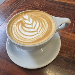 Latte - A latte with intricate latte art in a white ceramic cup on a wooden table, featuring a leaf pattern.