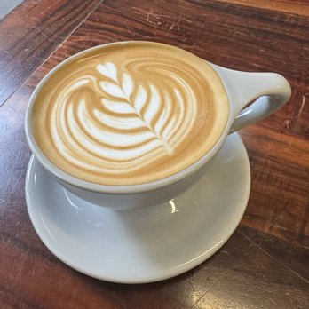 Theory Coffee Roasters - Latte - A latte with intricate latte art in a white ceramic cup on a wooden table, featuring a leaf pattern. - Coffee & Tea Near Me - Redding, California