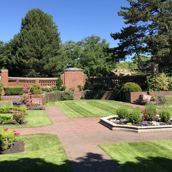 Lynch Park - Beautiful heritage garden and architecture. - A heritage garden featuring brick pathways, flower beds, and trimmed bushes, surrounded by tall trees and a brick wall. - Parks Near Me - Beverly, Massachusetts