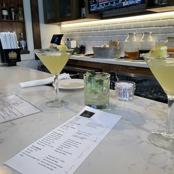 The Maine Catch - Cucumber Jalapeño Martini. - Two cucumber jalapeño martinis and a mojito are placed on a marble bar counter at The Maine Catch, with a menu nearby and bar supplies in the background. - Seafood Near Me - Ogunquit, Maine