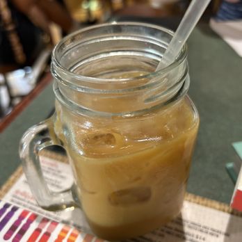 Maple + Bacon - Fluid coffee - A clear glass mason jar filled with iced coffee, ice cubes, and a straw is positioned on a restaurant table. The table surface is dark, and a colorful menu is partially visible underneath the jar. The background shows blurred figures of people and restaurant interior elements, indicating a casual dining setting. - Breakfast & Brunch Near Me - Valparaiso, Indiana