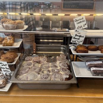Threefold Cafe - Pastries and baked goods. - The image shows a bakery display case filled with an assortment of baked items including cinnamon rolls, cookies, muffins, and biscuits. Handwritten labels identify items like cinnamon rolls, blueberry muffins, cheddar biscuits, and oatmeal cookies. The baked goods are arranged on white trays and plates, with some items wrapped in plastic. The background reflects a cafe setting with a person in a checkered shirt visible behind the counter. - Coffee & Tea Near Me - Chestnut Ridge, New York