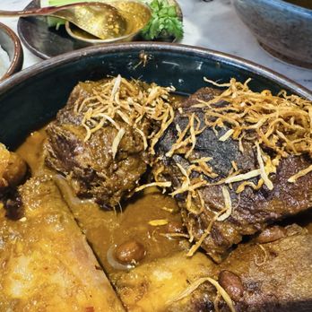 Sukh - A close-up of a bowl containing beef curry with shredded fried shallots on top, accompanied by rice and side dishes in the background. - Thai Near Me - Brooklyn, New York