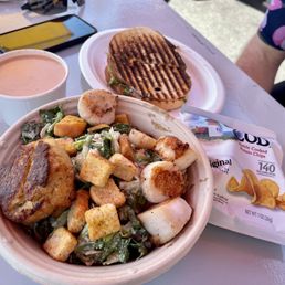 The image shows a meal with a bowl of salad containing croutons, grilled chicken, and leafy greens, a grilled sandwich with grill marks, a cup of creamy soup, and a packet of potato chips on a table. A smartphone and utensils are also visible on the table.
