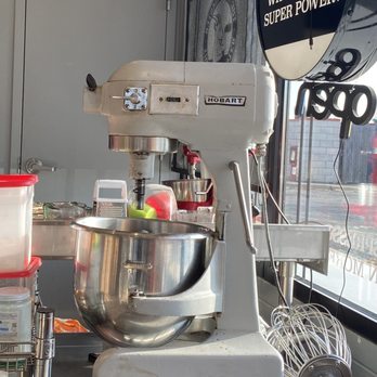 The Wild Roaster - Imagine the stories Big Bertha could tell if she could! - A white Hobart stand mixer with a stainless steel mixing bowl is positioned on a countertop in a kitchen or bakery setting. The mixer has control knobs on the front and is plugged into an electrical outlet. Nearby, there are various kitchen utensils, containers, and a window with a view outside. The scene suggests a professional or commercial food preparation environment. - Coffee & Tea Near Me - Deerfield, Illinois