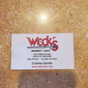 Weck's - A business card for Weck's restaurant in Santa Fe, NM, is shown on a textured, tan-colored surface. The card includes the restaurant's name, address at 2000 Cerrillos Rd., Santa Fe, NM 87505, and contact information such as phone number 505-471-9111, fax 505-471-9344, and website www.wecksinc.com. The card also features the name Cristina Castillo. The design includes the Weck's logo with a fork and knife graphic and the words 'Breakfast Lunch.' - Breakfast & Brunch Near Me - Santa Fe, New Mexico
