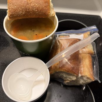 Panera Bread - Largest size Chicken Noodle Soup - A cup of chicken noodle soup with a bread roll on top, a sandwich in a plastic bag, and a small container of milk are placed on a black stovetop. - Soup Near Me - Cicero, New York