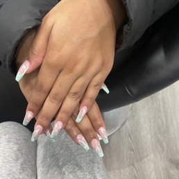 The image shows a pair of hands with manicured nails displaying a pastel pink and mint green design with white leaf-like patterns. The hands are resting on a person's lap, who is wearing light gray sweatpants. The background includes a black leather surface and part of a black jacket sleeve. The nails are long and well-shaped, with a glossy finish, and the design appears professionally done.
