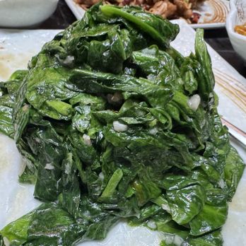 Juicy Dumplings - Pea Pod Stems - A large pile of cooked leafy greens, likely spinach or similar greens, is placed on a plate. The greens are dark green, slightly wilted, and appear to be seasoned or cooked with oil. In the background, there are other dishes, including noodles and possibly fried items, indicating a meal setting at an Asian restaurant. - Chinese Near Me - Euless, Texas