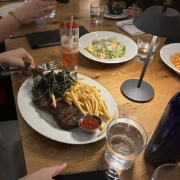 Steak Frites Dinner - A wooden dining table features a plate with a cooked steak topped with herbs, accompanied by French fries and a small container of ketchup. There are also salads, glasses of water, and colorful drinks with garnishes. Several people are seated around the table, some holding utensils or glasses, engaged in dining. The setting appears to be a casual restaurant with a lively atmosphere.