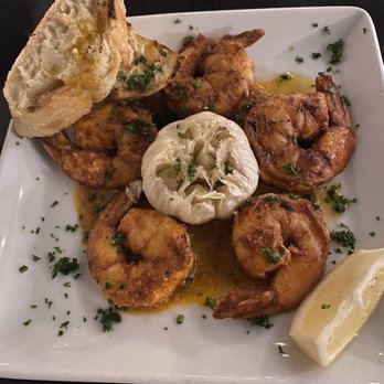 Debajo Tapas ABQ - Shrimp garlic head and aillijo (?) chili - The image shows a square white plate with several cooked shrimp garnished with chopped herbs, a lemon wedge on the side, a piece of garlic bread with herbs, and a halved artichoke in the center. The dish appears to be seasoned with herbs and oil, served on a dark surface. - Spanish Near Me - Albuquerque, New Mexico