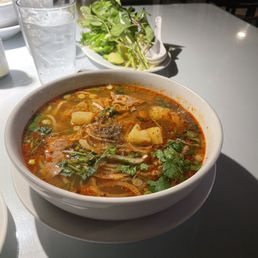 Rice Beef Soup - A bowl of spicy beef soup with vegetables, herbs, and broth is placed on a white table, with glasses of water and a plate of greens nearby.
