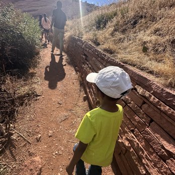 Red Rocks Park - Trail - A boy in a yellow shirt and white hat walks on a dirt trail, with a group of people ahead, surrounded by dry grass and a stone wall. - Parks Near Me - Morrison, Colorado