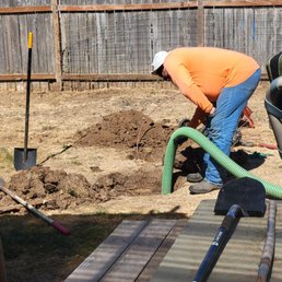 Gett'r done! - A person wearing an orange jacket and white hat is digging in a backyard, surrounded by tools like shovels and a flexible hose.