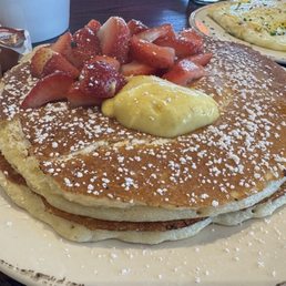 Lemon Ricotta Pancakes - A large white plate holds three thick, fluffy pancakes topped with sliced strawberries, a dollop of lemon curd, and a dusting of powdered sugar. The pancakes are stacked neatly, with the toppings arranged on top. In the background, there is a smaller plate with a fried egg and some condiments on a dark wooden table.