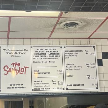The Sandlot - Menu - A menu board at The Sandlot in Park Ridge, Illinois, displays food and drink options such as milk, bottled water, shakes, and desserts like sundaes and cones. The menu includes prices and descriptions for items like the Two-N-Two burger, hot dogs, and ice cream treats. The board is mounted on a tiled wall with a ceiling vent visible above, and part of a staff member's cap with the business logo is seen at the bottom right. - Burgers Near Me - Park Ridge, Illinois