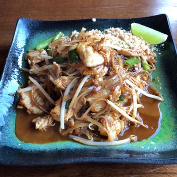 Ddee Thai - Pad thai with shrimp - A square black plate contains Pad Thai with shrimp, bean sprouts, and chopped green onions, topped with a sauce. A lime wedge is placed on the side of the plate. The dish is set on a wooden table in a restaurant setting. - Thai Near Me - Astoria, New York