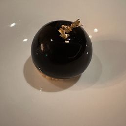 A glossy, black, spherical dessert with a smooth, reflective surface is presented on a white plate. The dessert is decorated with small pieces of gold leaf on top, creating an elegant appearance. The plate is plain and the background is neutral, emphasizing the dessert's sleek design.