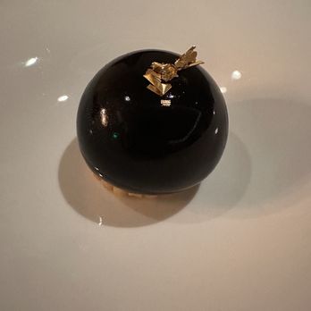 The Guest - A glossy, black, spherical dessert with a smooth, reflective surface is presented on a white plate. The dessert is decorated with small pieces of gold leaf on top, creating an elegant appearance. The plate is plain and the background is neutral, emphasizing the dessert's sleek design. - Modern European Near Me - Denver, Colorado