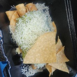 Sp#2 Carne Asada Burro, 3 Rolled Tacos - The image shows a black, rectangular takeout container filled with rolled tacos topped with shredded cheese. There are also tortilla chips placed alongside the tacos. The container appears to be made of foam or plastic, and the food is arranged in a simple manner, typical of takeout or delivery service from a Mexican restaurant or taqueria.
