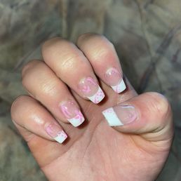 The image shows a close-up of a person's hand with five fingers visible, each with a French manicure featuring pink and white nail art. The nails have a clear base with white tips and pink decorative designs, including flowers and swirls. The background is blurred, possibly a stone or textured surface. The nails are well-groomed and the design is detailed and intricate.