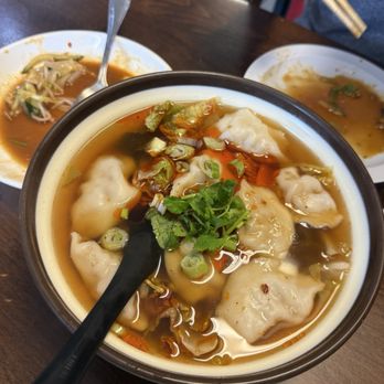 Wild Ginger - Ds1. Pork Dumplings in Soup - A bowl of wonton soup with dumplings, chopped green onions, cilantro, and vegetables on a wooden table, with side plates and chopsticks. - Chinese Near Me - Alameda, California