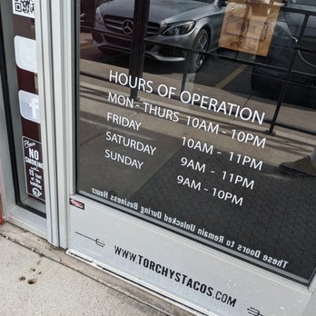Torchy's Tacos - Hours at Torchy's Tacos in Waco - The image features a glass storefront door with printed hours of operation for Torchy's Tacos in Waco, TX. The hours are listed for different days, with the website URL at the bottom. Reflections of parked cars and a person’s legs are visible in the glass, indicating an outdoor parking area nearby. - Tex-Mex Near Me - Waco, Texas