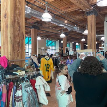 Retique - Nice interior! - The image shows the interior of a thrift store with a wooden ceiling and large wooden support beams. Multiple hanging lights illuminate the space, and large windows allow natural light to enter. Clothing racks filled with various garments, including a yellow sports jersey with the number 13, are visible. Several shoppers are browsing and interacting, including a woman in white and a person with curly hair taking a photo or adjusting their hair. The store has a spacious layout with a mix of organized clothing displays and open areas. - Thrift Stores Near Me - Milwaukee, Wisconsin