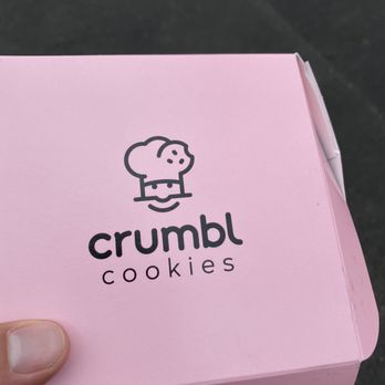 Crumbl - Delafield - A pink rectangular box from Crumbl Cookies displays a black logo of a chef's face with a hat and a smiling mouth. Below the logo, the text reads 'crumbl' in lowercase letters and 'cookies' underneath in smaller font. The box is held by a person's hand with a thumb visible in the lower left corner, and the background is a dark surface. - Bakeries Near Me - Delafield, Wisconsin