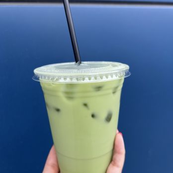 Cafe Select - Matcha latte - A hand holds a clear plastic cup filled with a green matcha latte containing ice cubes. The cup has a plastic lid with a black straw inserted. The background is a solid blue surface, possibly a wall or panel, providing a simple backdrop for the beverage. - Coffee & Tea Near Me - Fort Lee, New Jersey