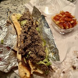Cheesesteak - A partially eaten cheesesteak sandwich with ground beef, lettuce, and bread, wrapped in foil, with a side of fries topped with ketchup in a plastic container.