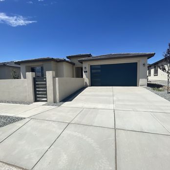 Beyond Inspections Service - The image shows a modern single-story house with a flat roof and a blue garage door. The concrete driveway leads directly to the garage, with a small gate and wall on the left side. The house has a neutral exterior color, and there are neighboring houses visible on both sides. The sky is clear and blue, indicating a sunny day. The front yard has minimal landscaping with a few small trees and gravel areas. - Home Inspectors Near Me - Albuquerque, New Mexico
