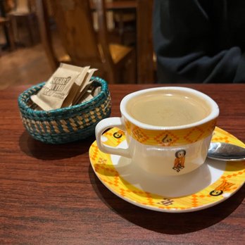 Lemat - Ethiopian Pot Coffee - A ceramic cup filled with Ethiopian coffee sits on a matching saucer with a yellow and orange pattern, featuring small illustrations. A metal spoon rests on the saucer, and a small woven basket containing sugar packets is placed nearby. The setting appears to be inside a restaurant with wooden furniture and chairs in the background, suggesting a cozy dining environment. - Ethiopian Near Me - Berkeley, California