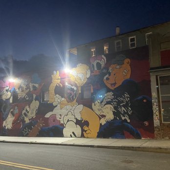 Electric Haze - The image shows a mural painted on the exterior wall of a building during nighttime. The mural features Disney characters such as Mickey Mouse, Minnie Mouse, and others, rendered in a colorful, cartoon style. The scene is illuminated by streetlights, creating reflections and glare on the wall. The building has multiple windows and a small storefront window on the right side, with a dark sky overhead. - Hookah Bars Near Me - Worcester, Massachusetts