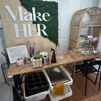 MAKE Collectives - Plant bar - A wooden table in a retail store features small potted plants, gardening tools, and decorative objects. Behind the table, a wall displays a sign reading 'Make HER' with a green textured background. To the right, a wicker display shelf holds additional items, and a small window is visible above. The space has a wooden floor and various storage containers underneath the table. - Vinyl Records Near Me - Long Beach, California