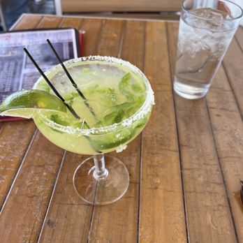 Thunderbird Bar & Grill - A margarita in a glass with salt rim, lime wedges, and two black straws on a wooden table, with a glass of water nearby. - American Near Me - Santa Fe, New Mexico