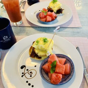 Blue Shutters Inn and Studios - Gourmet Breakfast - A breakfast scene at Blue Shutters Inn and Studios in Ogunquit, ME, showing eggs benedict topped with green onions on a white plate, accompanied by a bowl of cubed watermelon with a mint garnish, and a Bloody Mary cocktail garnished with pickles and lemon. The table has pink placemats, and black mugs are visible in the background. - Hotels Near Me - Ogunquit, Maine