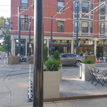 Cobblestone OTR - The image depicts an outdoor seating area of a restaurant or cafe situated on a city street. There are tables and chairs arranged on a wooden deck, with large planters containing greenery. The background features a multi-story building with bay windows and a storefront with visible patrons inside. Street elements such as traffic cones, parked cars, and overhead tram wires are also present, indicating an urban environment. - Cocktail Bars Near Me - Cincinnati, Ohio